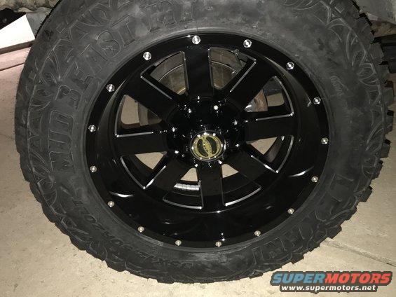 Black aftermarket wheel with chrome accents and a mud-terrain tire. Center cap has a gold logo