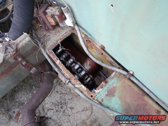Rusty mechanical linkage with a black chain inside a metal housing, with hoses and wires attached