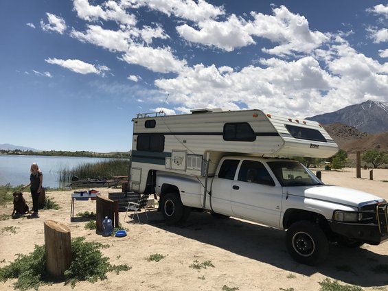 White pickup truck with a camper shell parked on a dirt lot near a lake. The truck has a bull bar and dual rear wheels