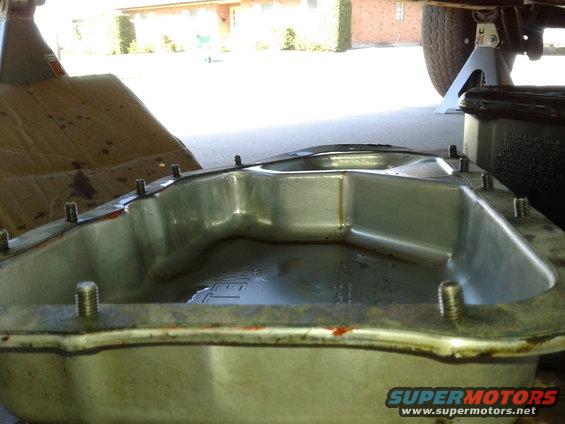 Oil pan with bolts removed, sitting on the ground. A car tire and jack stand are visible in the background