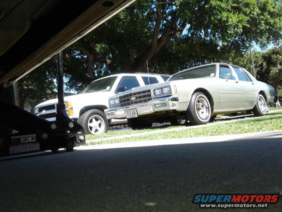 Silver 1980s Chevrolet Caprice sedan parked next to a white SUV. A car jack is visible in the foreground