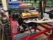 1964 Ford Galaxie - A V8 engine with gold valve covers and custom headers is mounted on a red stand, with parts of two pickup trucks visible in the background.
