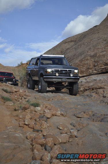 Dark gray Ford Bronco climbing a rocky trail with a red Jeep behind it