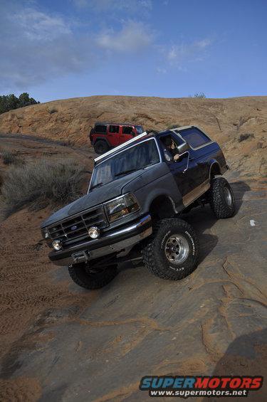 Dark gray Ford Bronco climbing rocks, red Jeep in background