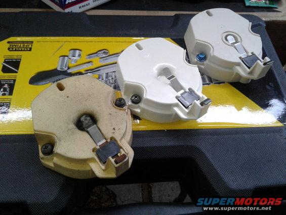 Three distributor caps, one tan and two white, rest on a black surface with a yellow tool packaging behind them