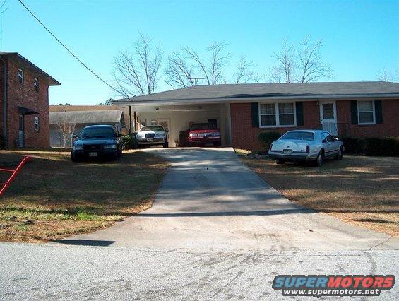 Four cars parked in front of a house and carport. One is a dark sedan, two are lighter sedans, and one is a red sedan
