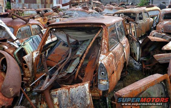 Junkyard scene with many heavily rusted cars piled together. Focus on the rear of a station wagon with its interior exposed