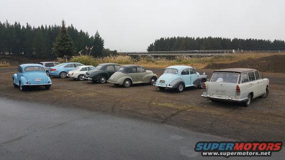 Several vintage cars parked in a lot, including a light blue sedan, a dark sedan, a light gray sedan, and a light-colored station wagon