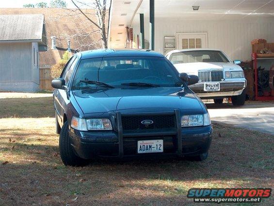 Black 2001 Ford Crown Victoria with a push bar and license plate ADAM-12 parked on grass