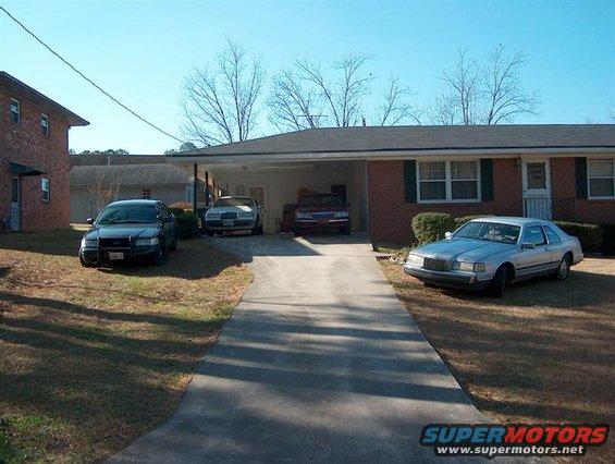 Several cars parked in front of a house, including a police-style sedan and a silver coupe