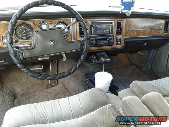 1984 Buick Electra interior with wood grain trim, leather-wrapped steering wheel, and velour seats. A white cup sits in the center console