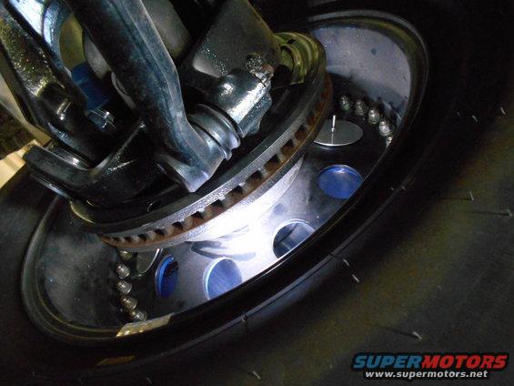 Close-up of a vehicle's wheel hub, brake rotor, and suspension components. The wheel has a polished metal rim with visible lug nuts and blue accents