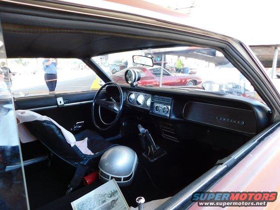 Interior view of a 1968 Mercury Cougar with racing bucket seats, floor shifter, and a tachometer. Another red car is visible outside