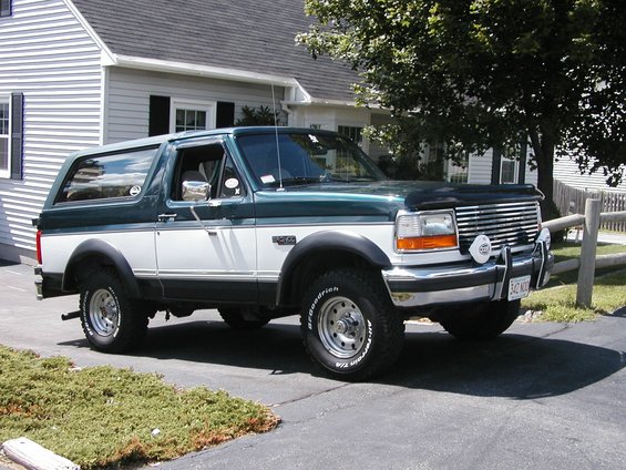 Green and white Ford Bronco parked in driveway, front quarter view. Has aftermarket lights on the bumper