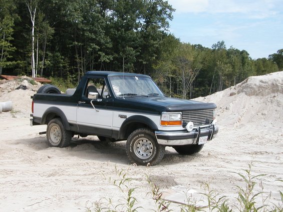 Two-tone dark green and white Ford Bronco with a spare tire on the back, parked on sandy terrain