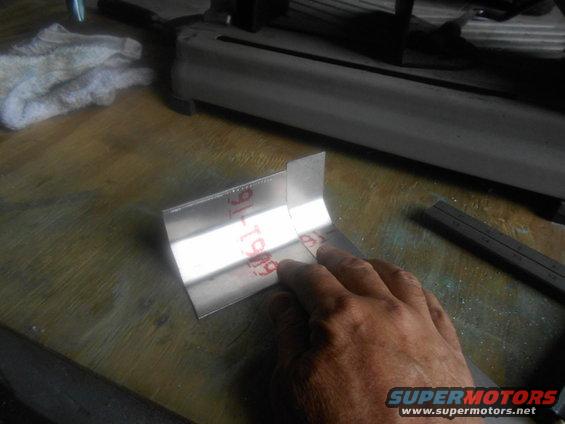 Metal bracket with red markings '91-T909' held by a hand over a wooden surface near a cutting tool