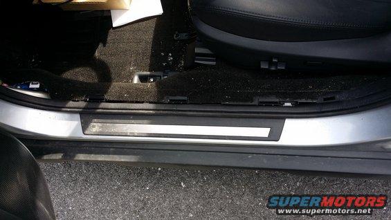 Car interior door sill with black carpet and a silver trim piece. A black leather seat is visible