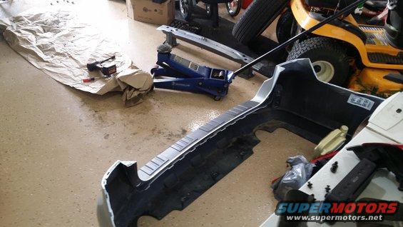 Car bumper and blue hydraulic jack on garage floor. Lawn mower in background