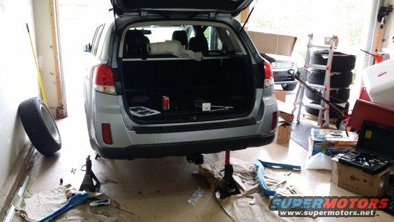 Rear of a silver Subaru Outback wagon lifted on jack stands in a garage. A tire is leaning against the wall. Tools and parts are on the floor