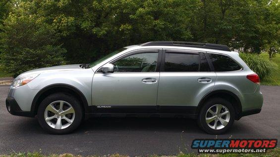 Side view of a silver Subaru Outback wagon parked on pavement with trees in the background. The word 'OUTBACK' is visible on the lower door trim
