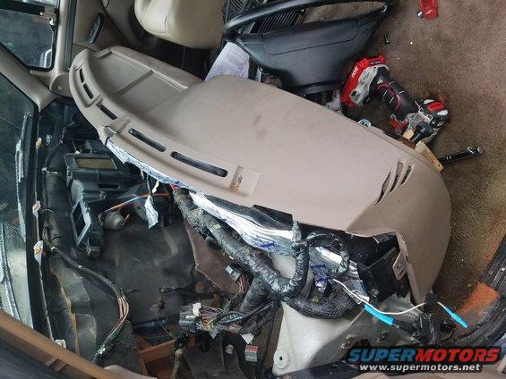Disassembled tan dashboard with exposed wiring and insulation. A red and black power drill rests on the dashboard