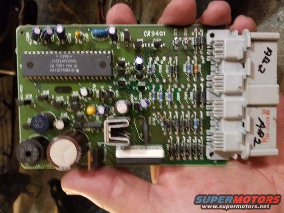 Green circuit board with a large black chip and white connectors, held by a hand