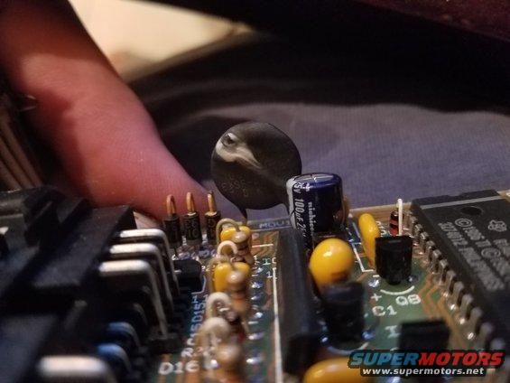 Circuit board with capacitors, resistors, and a black component with a white stripe. A finger is visible on the left
