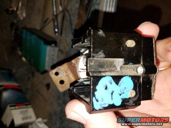 Close-up of a black automotive electrical component with blue markings and a rusty metal bracket