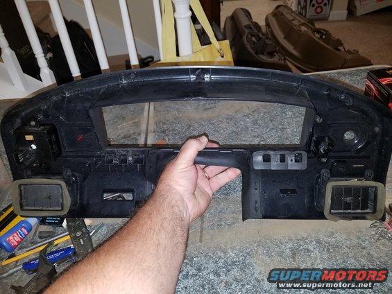 Black dashboard trim panel held by a hand, showing air vents and mounting points