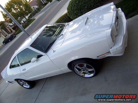 White 1978 Ford Mustang II coupe parked on a concrete driveway. Another dark car is visible on the street in the background