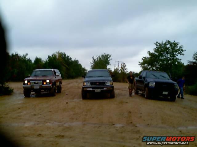 Three SUVs and trucks on a dirt road under a cloudy sky. The leftmost vehicle is a red SUV, possibly a 1997 Ford Bronco. A dark SUV is in the middle, and a dark pickup truck is on the right
