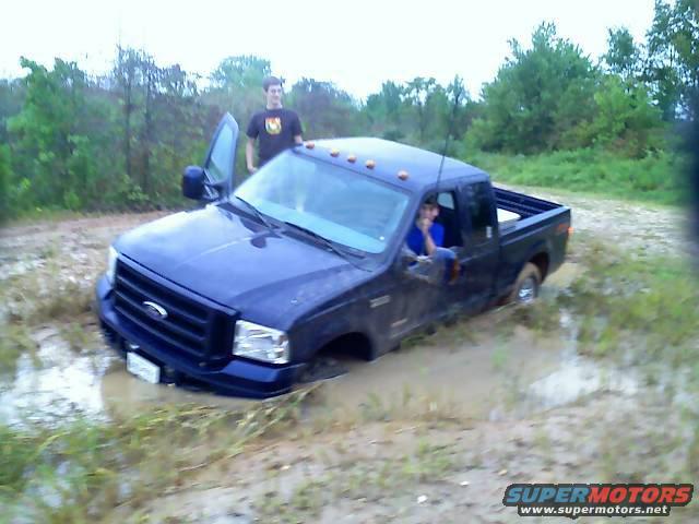 Blue pickup truck stuck in mud, front view. Driver in cab, person standing by open door