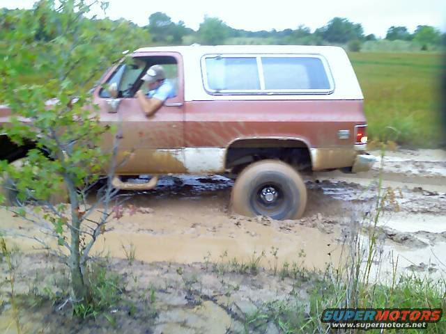 Red and white SUV with a white camper shell stuck in thick mud, driver visible