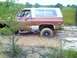 1989 Ford Bronco - A red and white SUV is stuck deep in mud, with its driver visible behind the wheel.