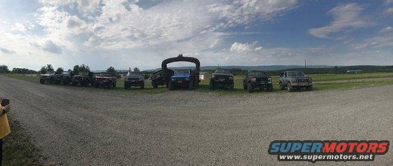 Several SUVs and trucks parked in a line on a gravel road, with a large archway structure in the background