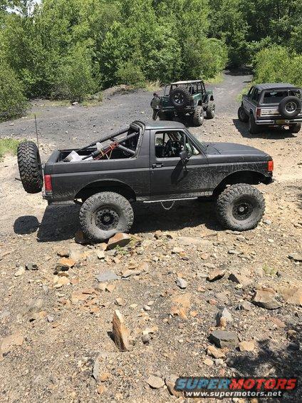 Modified 1989 Ford Bronco with external roll cage and spare tire on rear, parked on rocky terrain with other off-road vehicles