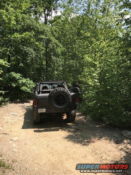 Black Ford Bronco with spare tire on back, parked on dirt road in woods