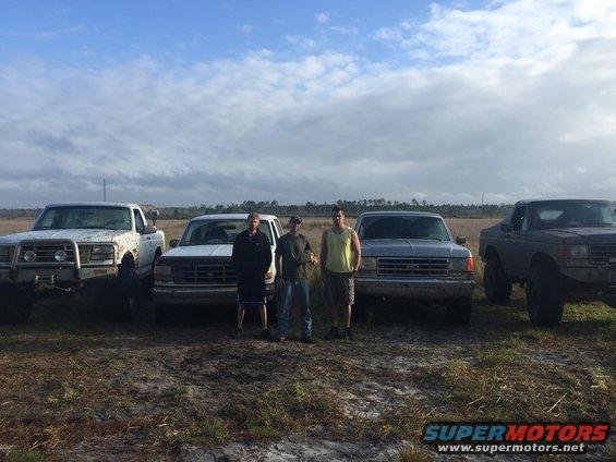 Four pickup trucks parked in a field with three men standing in front of them. Trucks appear to be from the late 1980s or early 1990s