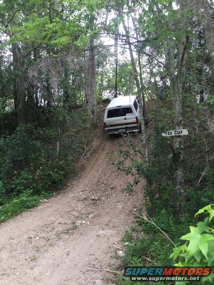 1989 Ford Bronco driving up a steep dirt hill in a wooded area. Sign reads 'TEA CUP'