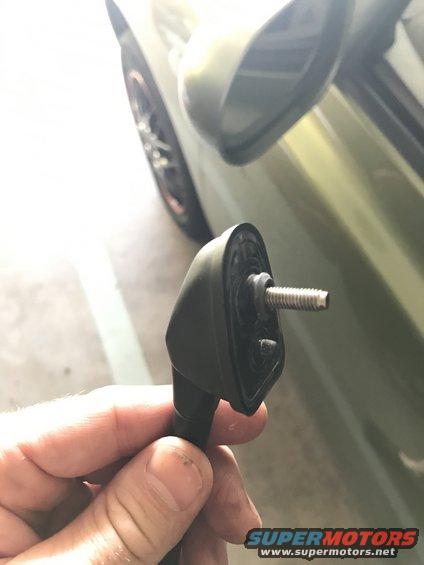 Hand holding black car side mirror mounting bracket with screw, green car door and wheel visible