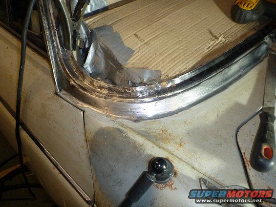 Close-up of a car's body panel undergoing repair, showing rusted metal, chrome trim, and tools like clamps and a file