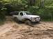 1989 Ford Bronco - A white Ford Bronco navigates a challenging muddy path in a wooded area, with another vehicle following behind.