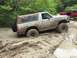 1989 Ford Bronco - Two Ford Broncos navigate a muddy off-road trail, with the white Bronco partially submerged in thick mud.