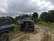 1989 Ford Bronco - Two off-road vehicles are parked on a dirt path with trees and cloudy skies in the background.