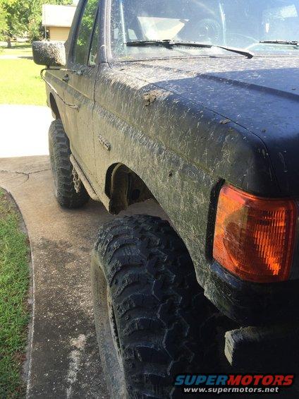 Mud-covered black 1987 Ford Bronco, front passenger side, angled from the ground up, showing large off-road tires and orange turn signal