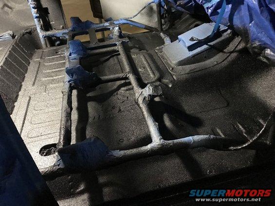 Vehicle undercarriage with frame rails and floor pan coated in black protective material, some parts masked with blue tape