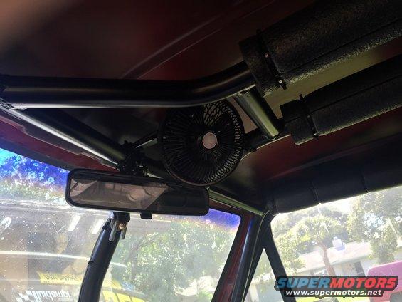 Interior view of a vehicle showing a black fan mounted above a rearview mirror, with a roll cage visible