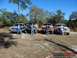 1989 Ford Bronco - Four trucks and SUVs are parked in a grassy area with trees in the background under a clear blue sky.