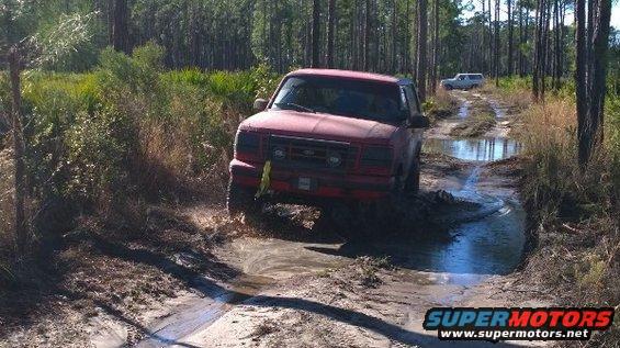 1989 Ford Bronco driving through a muddy trail with another SUV in the background