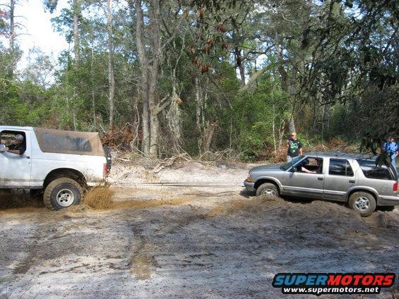 White SUV with soft top stuck in mud next to a silver SUV in a wooded area
